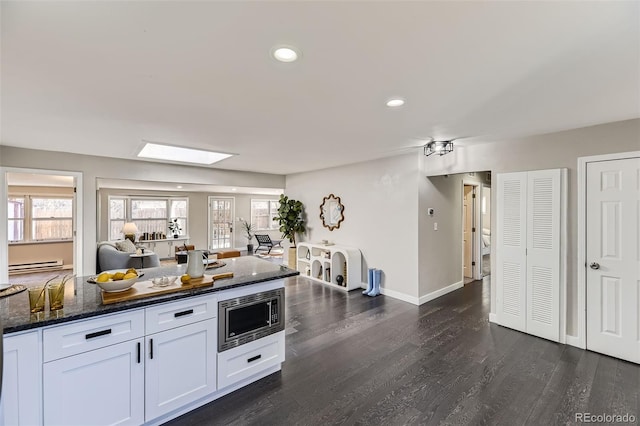 kitchen with dark wood finished floors, stainless steel microwave, baseboard heating, white cabinetry, and dark stone countertops