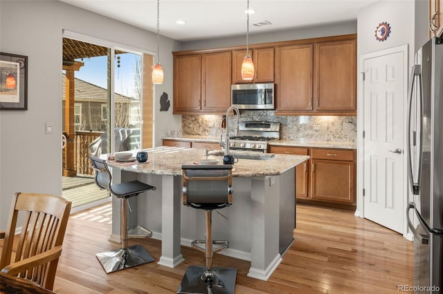 kitchen with an island with sink, brown cabinets, backsplash, and appliances with stainless steel finishes