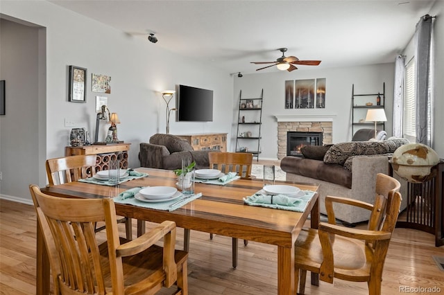 dining space featuring baseboards, a stone fireplace, light wood-type flooring, and a ceiling fan