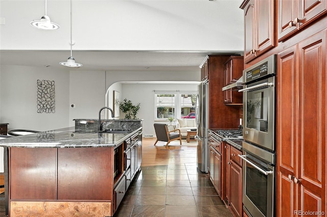 3608 S Newport Way, Denver, CO 80237 - kitchen featuring appliances with stainless steel finishes, dark stone counters, pendant lighting, an island with sink, and arched walkways