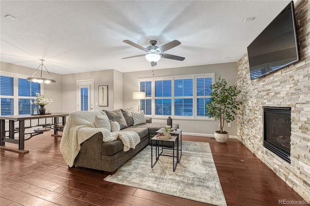 living room featuring dark wood-style floors, ceiling fan, a stone fireplace, and a textured ceiling