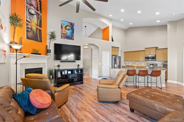 living room with a tile fireplace, light hardwood / wood-style floors, high vaulted ceiling, sink, and ceiling fan with notable chandelier