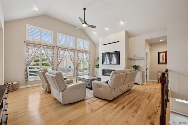 living area with a ceiling fan, a glass covered fireplace, light wood-style floors, high vaulted ceiling, and baseboards