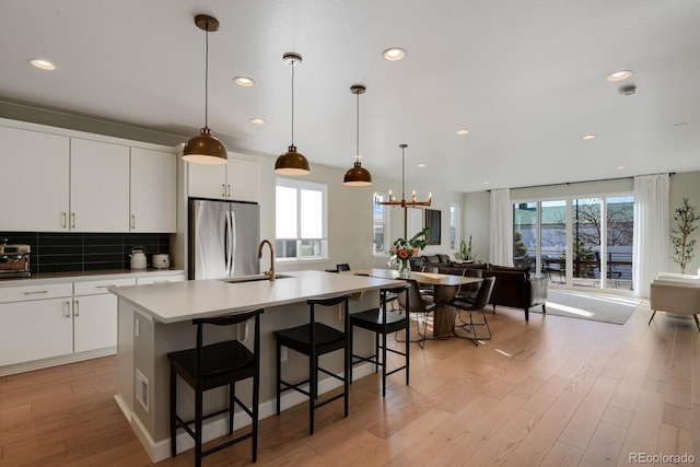 kitchen with tasteful backsplash, decorative light fixtures, a center island with sink, stainless steel refrigerator, and white cabinets