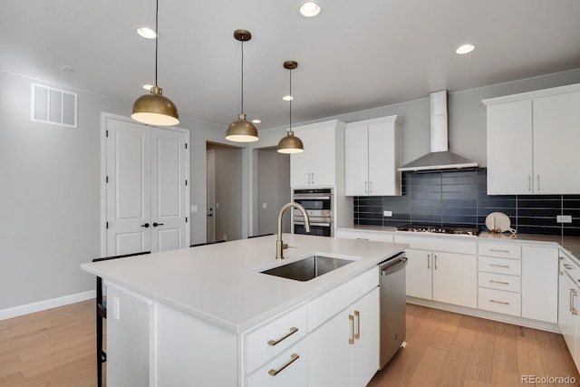 kitchen with wall chimney exhaust hood, white cabinetry, a center island with sink, appliances with stainless steel finishes, and pendant lighting