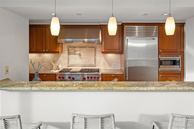 kitchen with light stone counters, wall chimney exhaust hood, brown cabinetry, decorative backsplash, and built in appliances