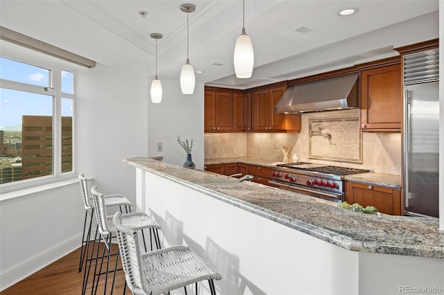 kitchen with high quality appliances, wall chimney exhaust hood, decorative light fixtures, light stone counters, and a breakfast bar area