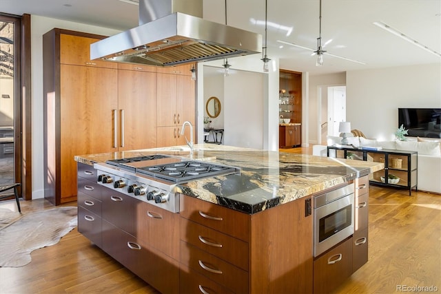 kitchen featuring stone countertops, a large island with sink, island exhaust hood, stainless steel appliances, and light hardwood / wood-style flooring