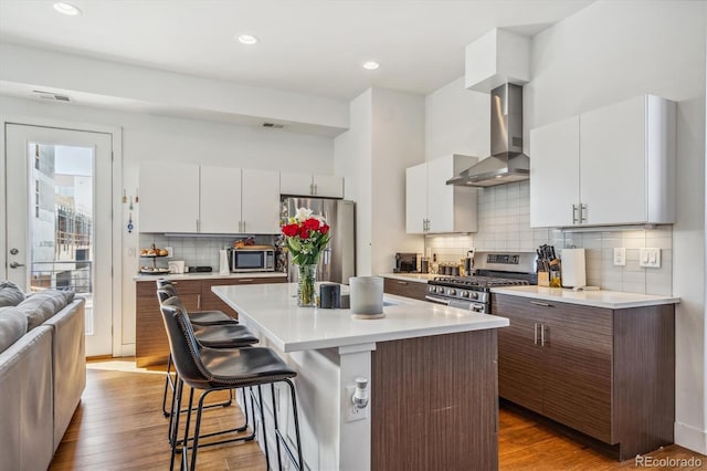 kitchen featuring wall chimney exhaust hood, wood-type flooring, a kitchen island, a breakfast bar, and appliances with stainless steel finishes