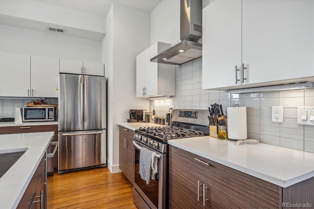 kitchen with appliances with stainless steel finishes, light wood-type flooring, white cabinetry, wall chimney range hood, and backsplash