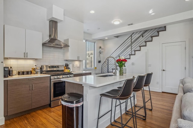 kitchen featuring stainless steel gas range oven, wall chimney exhaust hood, a kitchen island with sink, white cabinetry, and sink