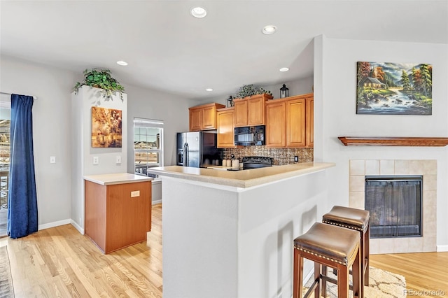 kitchen featuring decorative backsplash, a tiled fireplace, light wood-style flooring, a peninsula, and black appliances