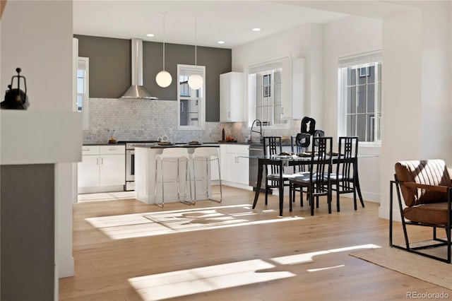 kitchen with wall chimney range hood, light hardwood / wood-style flooring, stainless steel stove, white cabinetry, and hanging light fixtures