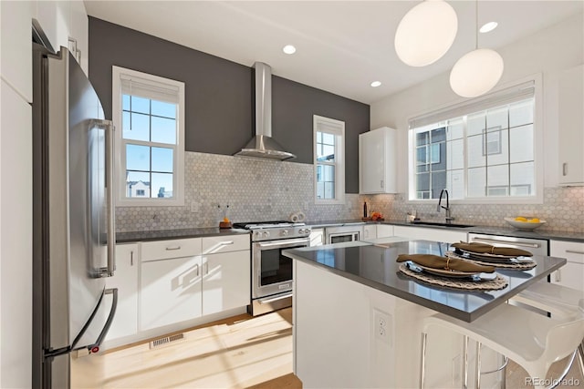 kitchen with white cabinetry, stainless steel appliances, hanging light fixtures, and wall chimney range hood