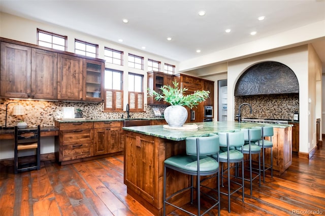 kitchen with light stone countertops, decorative backsplash, dark hardwood / wood-style flooring, a breakfast bar, and a center island with sink