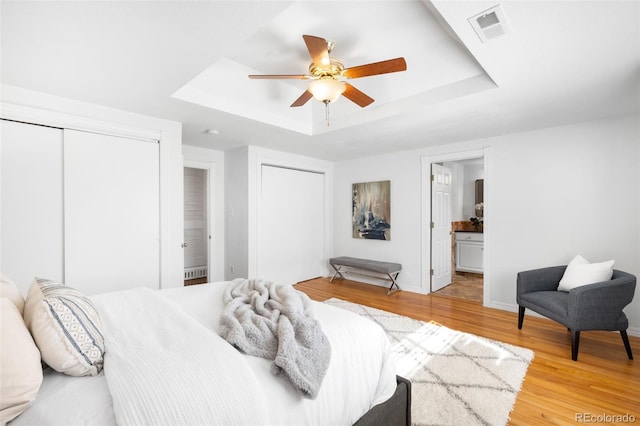 bedroom featuring light wood finished floors, visible vents, two closets, ceiling fan, and a tray ceiling