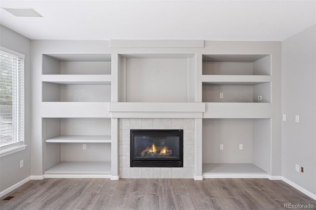 unfurnished living room featuring wood finished floors, built in shelves, a tiled fireplace, baseboards, and visible vents