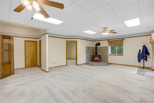 basement with a wood stove, a paneled ceiling, ceiling fan, and light carpet