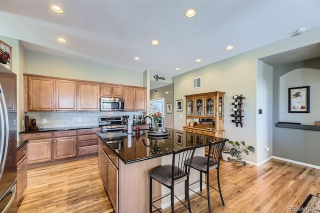 kitchen with light wood-type flooring, backsplash, dark stone counters, stainless steel appliances, and a kitchen island with sink