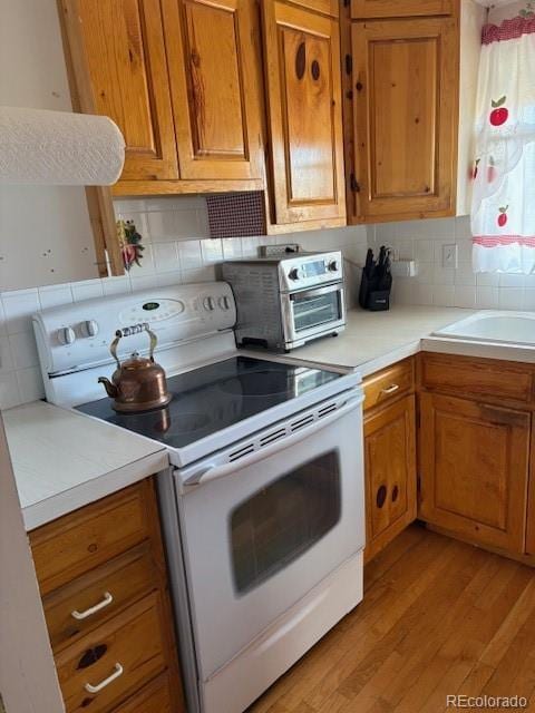 kitchen with white electric range oven, brown cabinetry, light countertops, light wood-type flooring, and backsplash