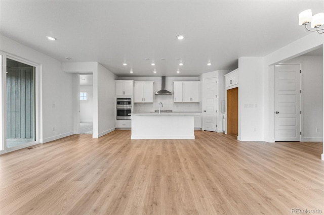 kitchen with white cabinetry, wall chimney range hood, decorative backsplash, light wood-type flooring, and a center island with sink