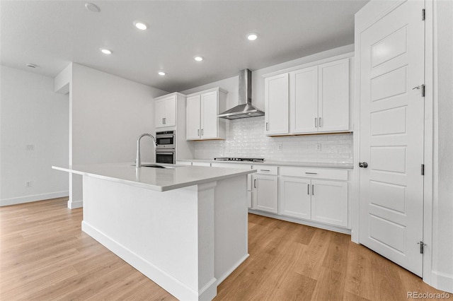 kitchen with white cabinetry, wall chimney range hood, a center island with sink, and light wood-type flooring