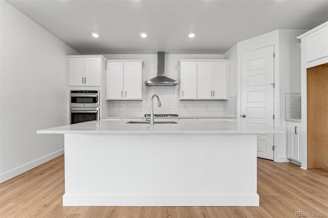 kitchen with white cabinetry, wall chimney range hood, double oven, light hardwood / wood-style floors, and a kitchen island with sink