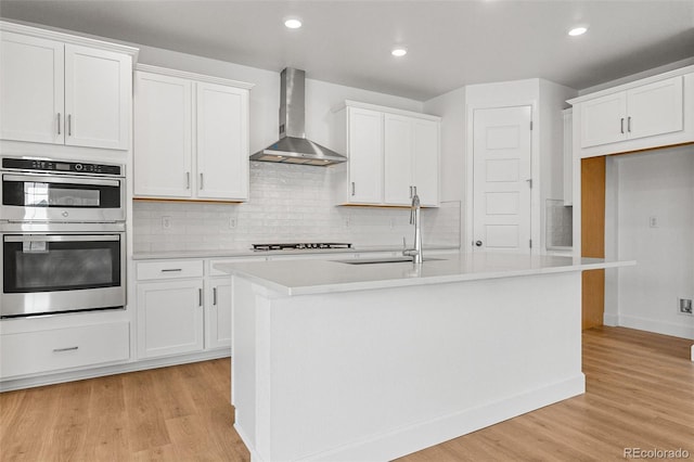 kitchen featuring sink, white cabinetry, a kitchen island with sink, stainless steel double oven, and wall chimney exhaust hood