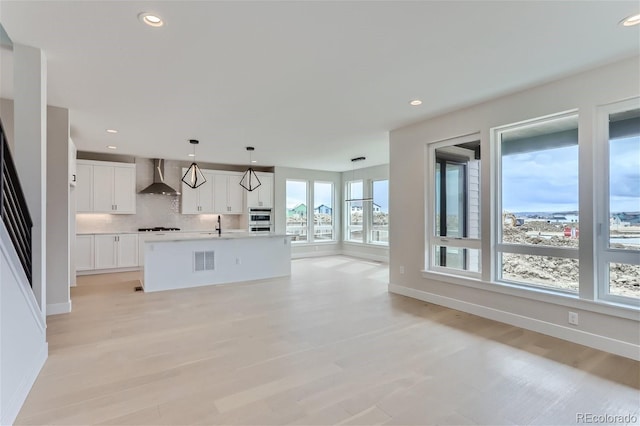 kitchen with wall chimney exhaust hood, pendant lighting, a kitchen island with sink, decorative backsplash, and white cabinets