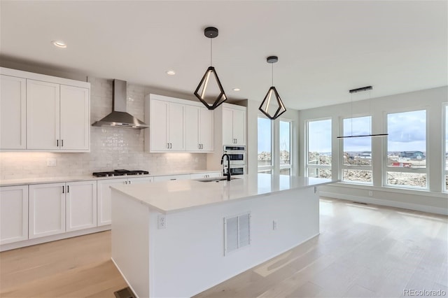 kitchen with pendant lighting, sink, white cabinets, a center island with sink, and wall chimney exhaust hood