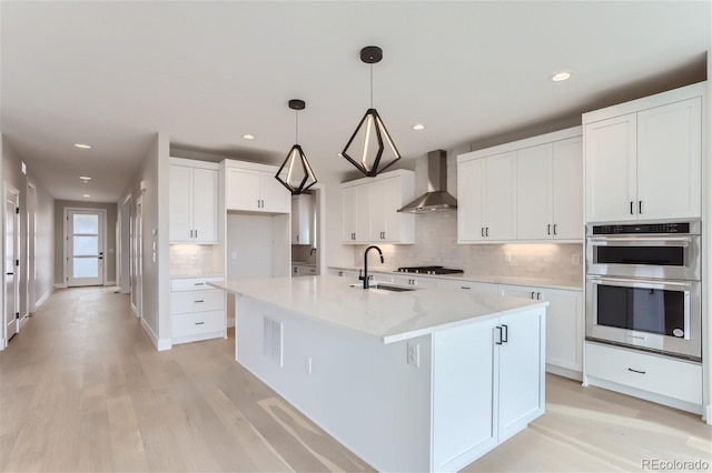 kitchen featuring wall chimney range hood, sink, a kitchen island with sink, white cabinetry, and hanging light fixtures