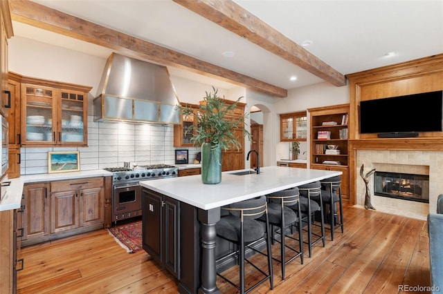 kitchen featuring beam ceiling, an island with sink, range hood, arched walkways, and designer stove