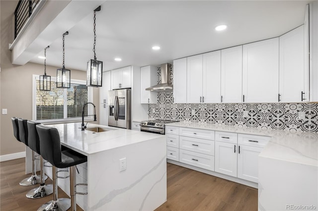 kitchen with appliances with stainless steel finishes, wall chimney range hood, sink, white cabinetry, and an island with sink