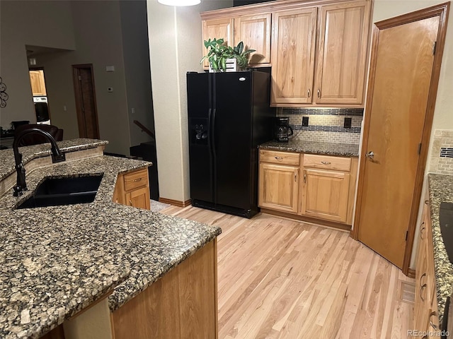 kitchen with light wood-type flooring, a sink, backsplash, dark stone counters, and black refrigerator with ice dispenser