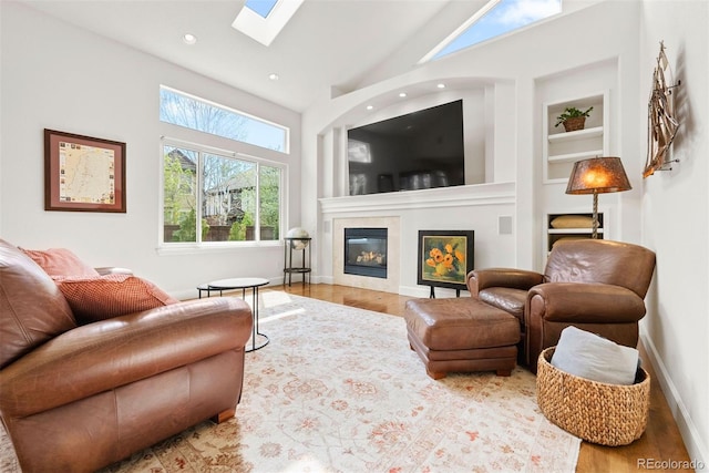 living area featuring a glass covered fireplace, wood finished floors, a high ceiling, built in shelves, and a skylight