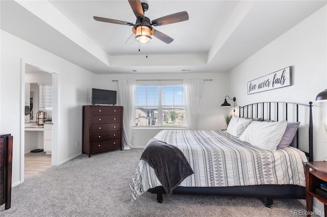 bedroom featuring a tray ceiling, ensuite bath, light colored carpet, and ceiling fan