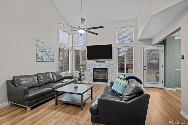 living room featuring ceiling fan, a tile fireplace, light wood-style flooring, a high ceiling, and baseboards