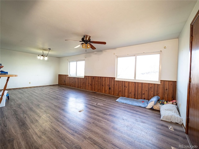 spare room featuring dark wood finished floors, ceiling fan with notable chandelier, wood walls, and wainscoting