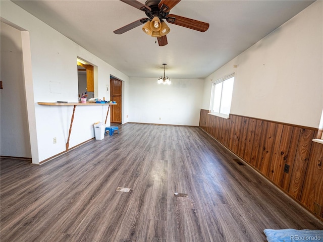 unfurnished living room with visible vents, wooden walls, a wainscoted wall, ceiling fan with notable chandelier, and dark wood-style flooring