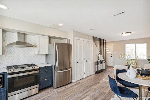 kitchen with appliances with stainless steel finishes, tasteful backsplash, white cabinetry, wall chimney range hood, and light wood-type flooring