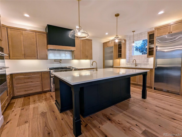 kitchen with high quality appliances, a center island with sink, hanging light fixtures, light hardwood / wood-style flooring, and custom range hood