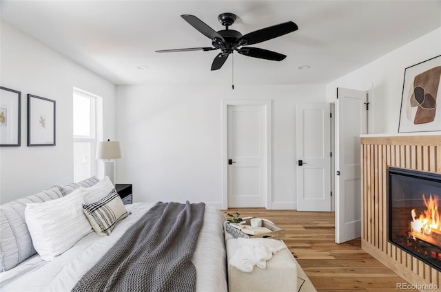 bedroom featuring a ceiling fan, a glass covered fireplace, baseboards, and wood finished floors