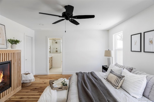 bedroom with a warm lit fireplace, wood finished floors, a ceiling fan, baseboards, and ensuite bath