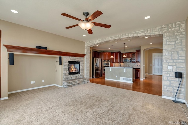 unfurnished living room featuring ceiling fan, arched walkways, a stone fireplace, dark colored carpet, and visible vents