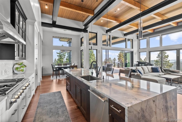 kitchen with wood ceiling, light stone counters, an island with sink, a towering ceiling, and appliances with stainless steel finishes