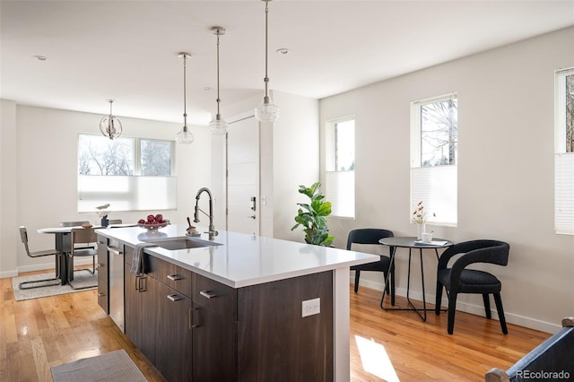 kitchen with light wood-type flooring, a center island with sink, dark brown cabinetry, light countertops, and hanging light fixtures