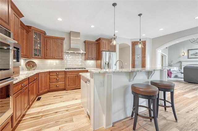 9121 Sunshine Meadow Pl, Parker, CO 80134 - kitchen with wall chimney range hood, stainless steel appliances, arched walkways, light wood-style floors, and brown cabinets