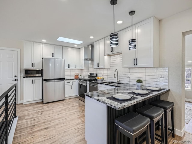 kitchen featuring wall chimney exhaust hood, appliances with stainless steel finishes, a peninsula, light wood-style floors, and a sink