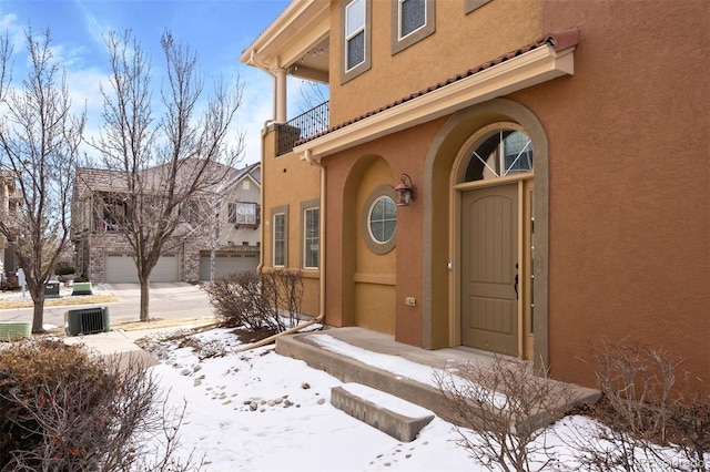 snow covered property entrance featuring a garage, a balcony, and stucco siding