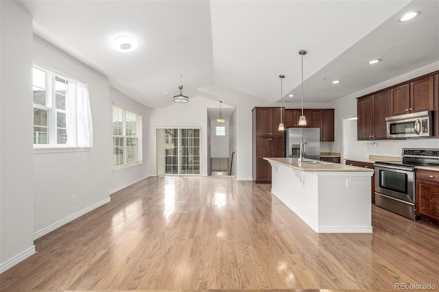 kitchen featuring stainless steel appliances, an island with sink, pendant lighting, vaulted ceiling, and light wood-type flooring